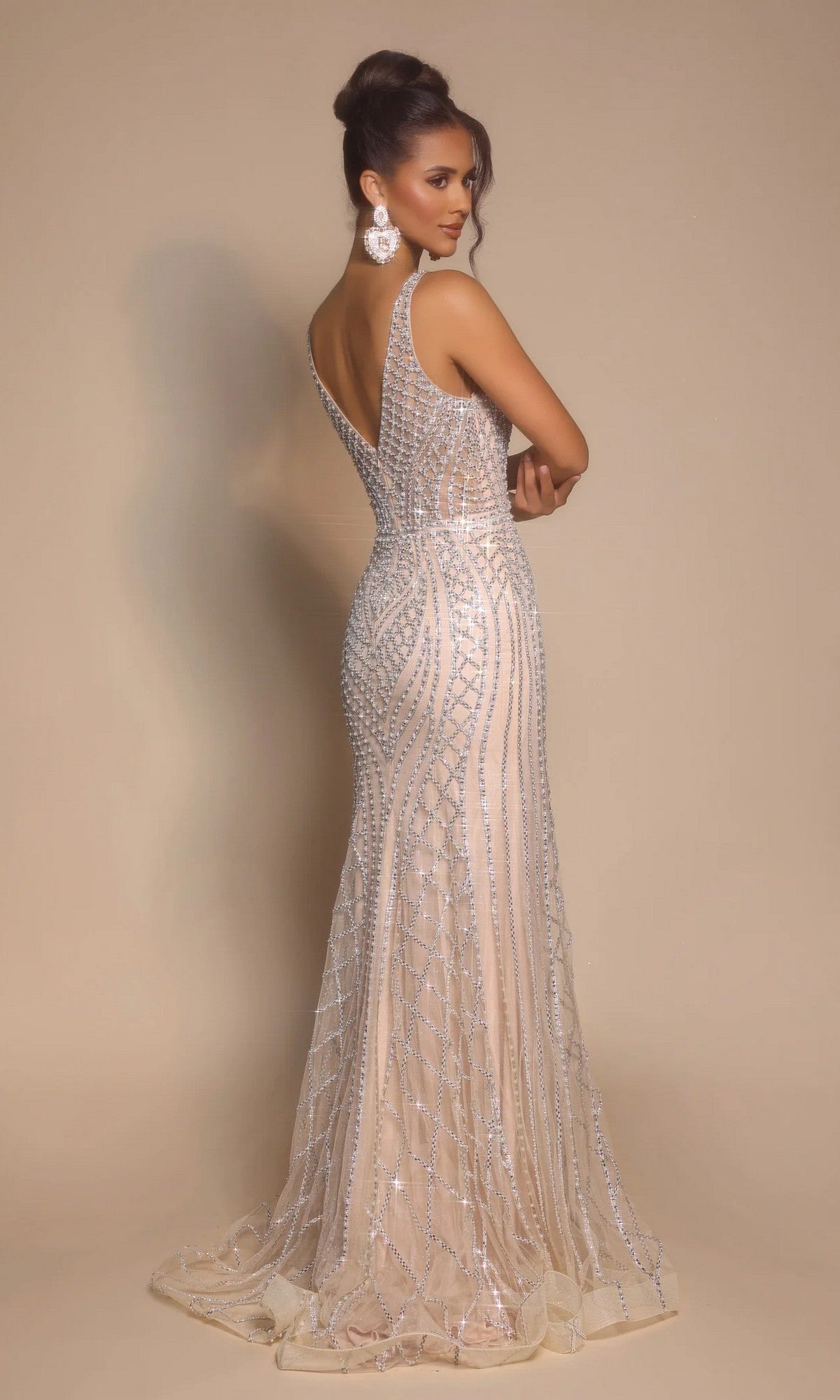 A woman stands with her back turned, looking over her shoulder, wearing the Portia and Scarlett PS26089 long formal dress—an embellished, sleeveless gown with silver details—against a neutral background.